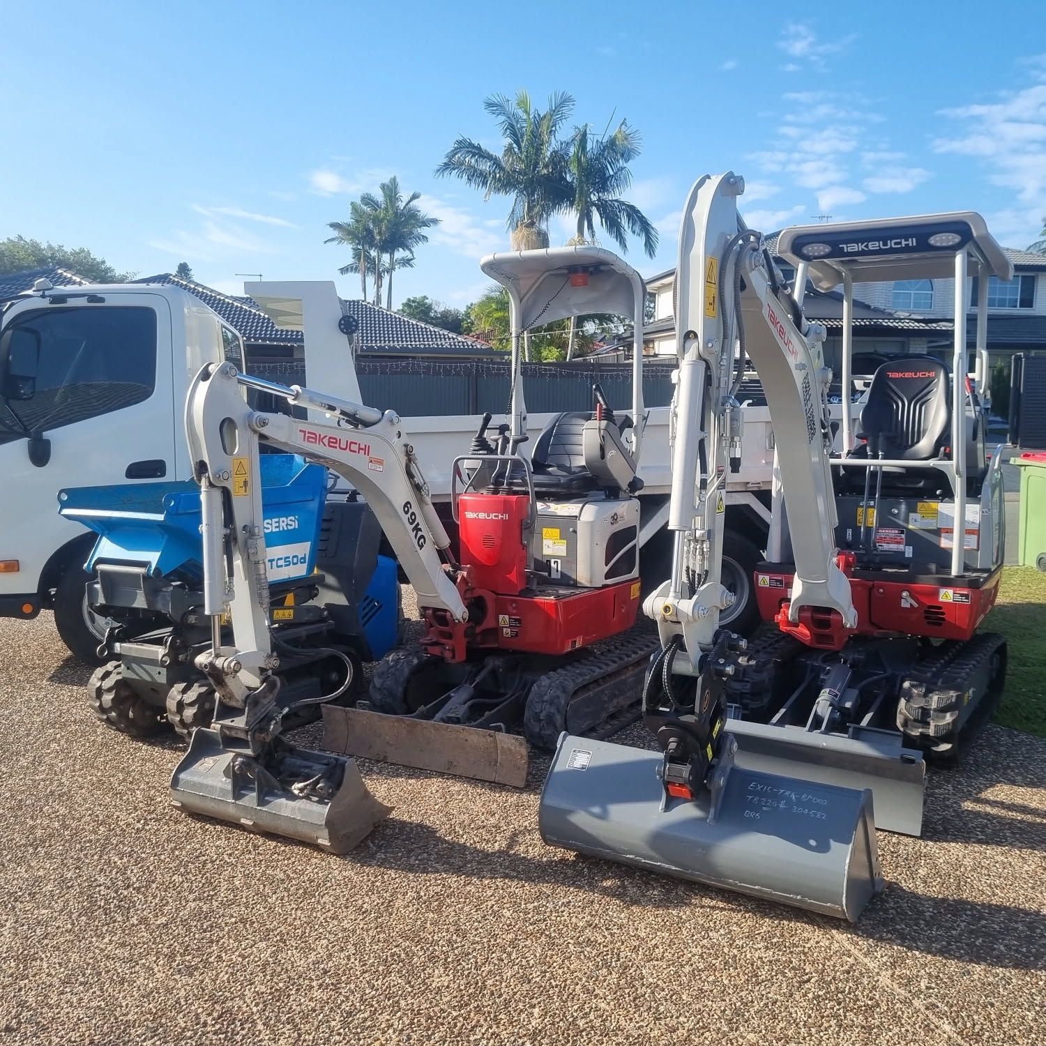 Two small excavators and a truck on gravel in front of a building with palm trees— Coastline Earthworks in Helensvale, QLD