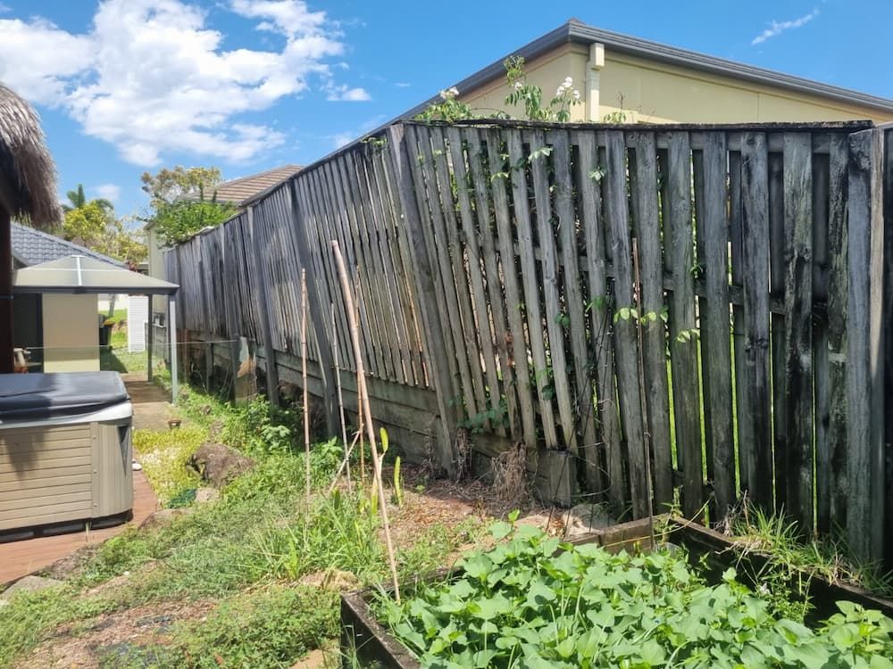 Worn, leaning wooden fence in a backyard, beside a garden with plants on a sunny day. — Coastline Earthworks in Helensvale, QLD