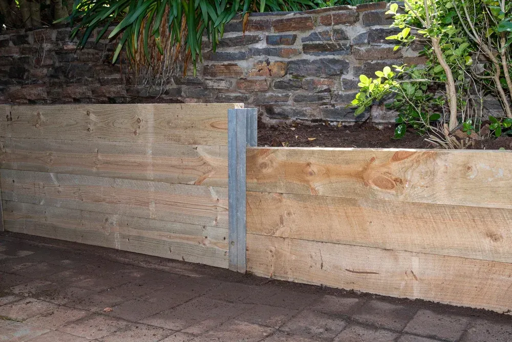 Wooden retaining wall with a stone wall backdrop, supported by a metal post. — Coastline Earthworks in Helensvale, QLD