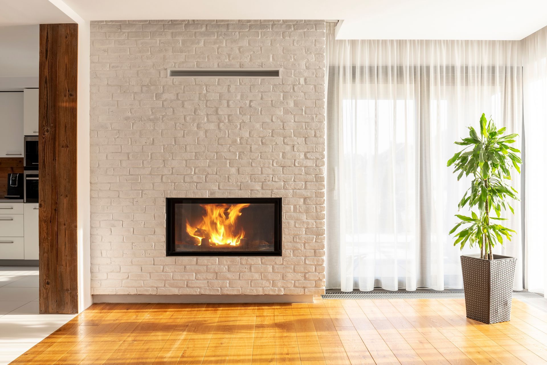 Fireplace with flames in a white brick wall, beside a plant in a pot and sheer curtains.