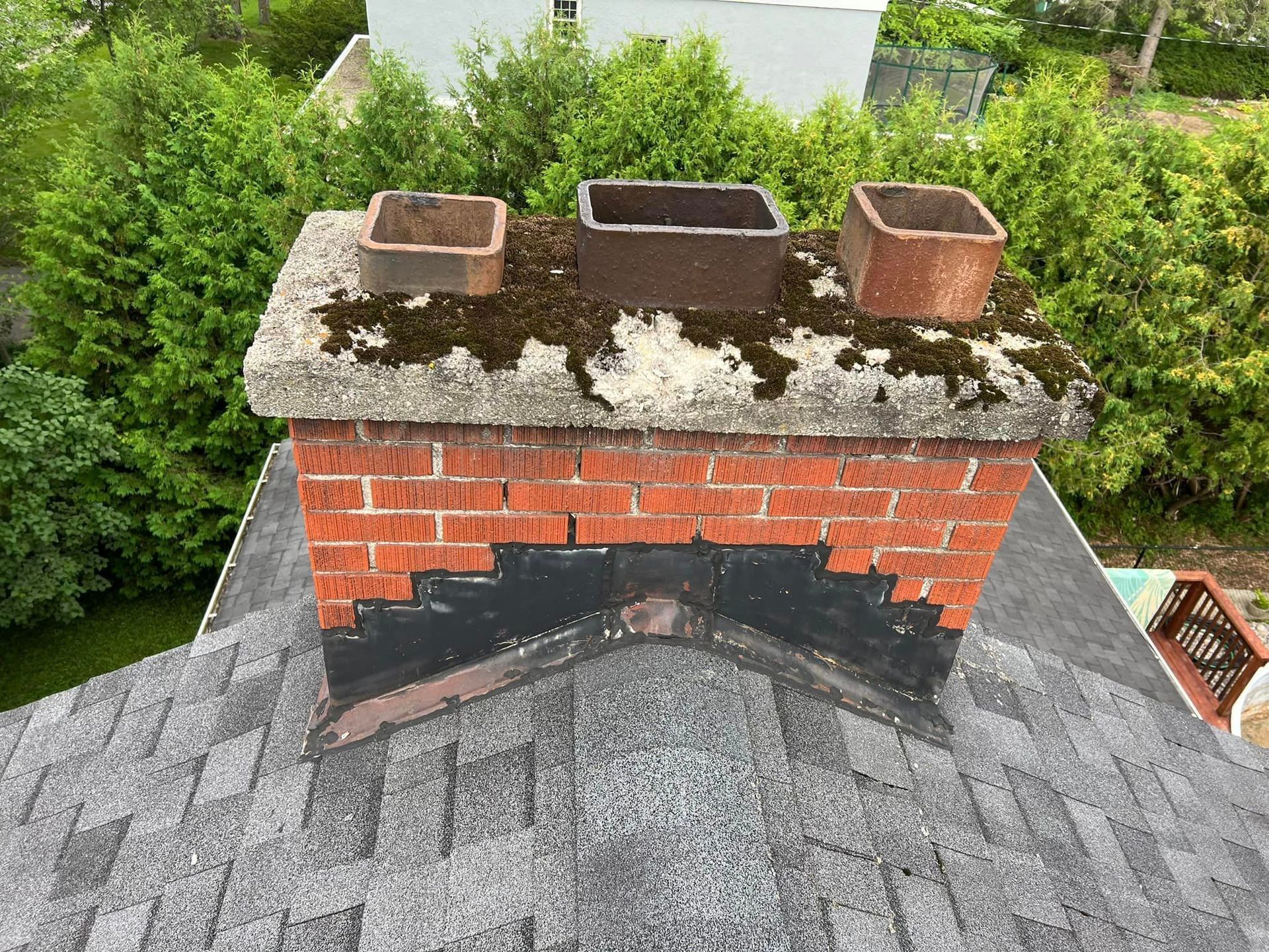 Brick chimney on a gray shingle roof with black discoloration, topped with three rusty metal flues.