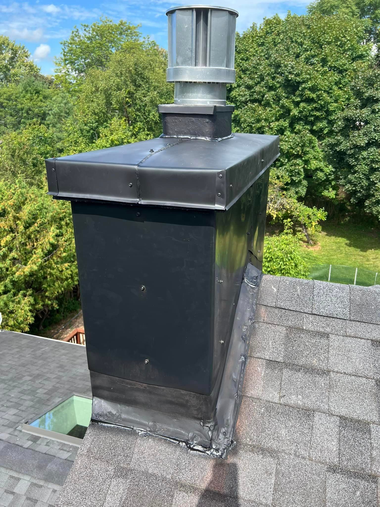 Black chimney with metal cap on a shingled roof, surrounded by green trees and a blue sky.