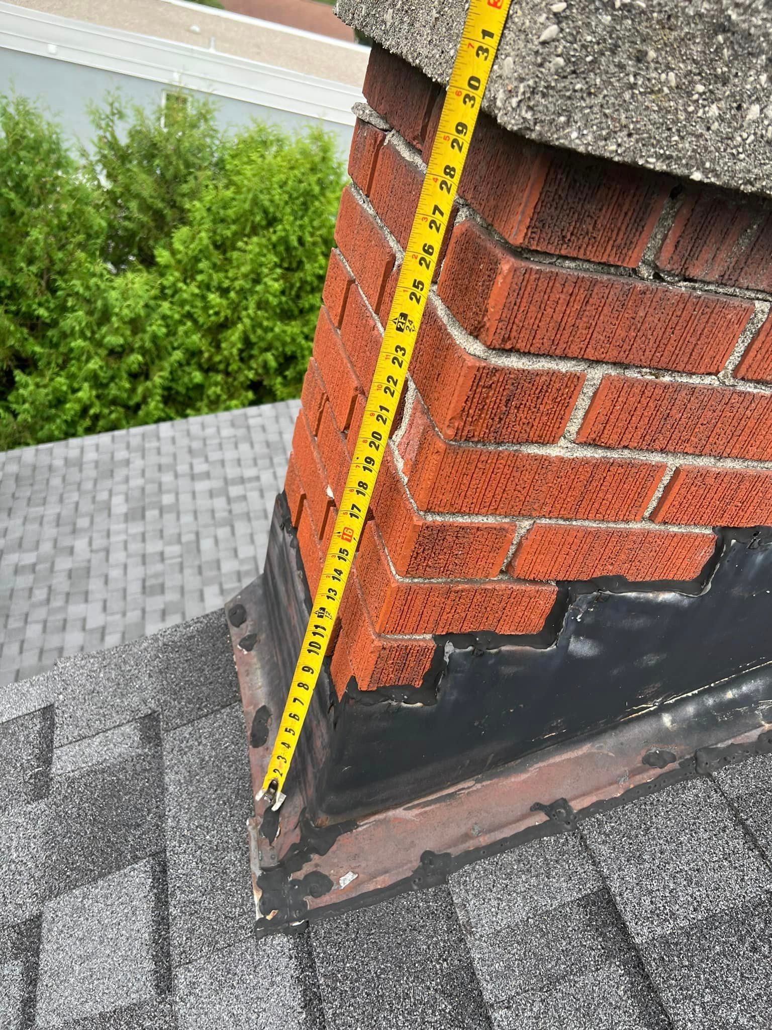 A chimney made of red brick on a gray shingle roof. A measuring tape shows its height.