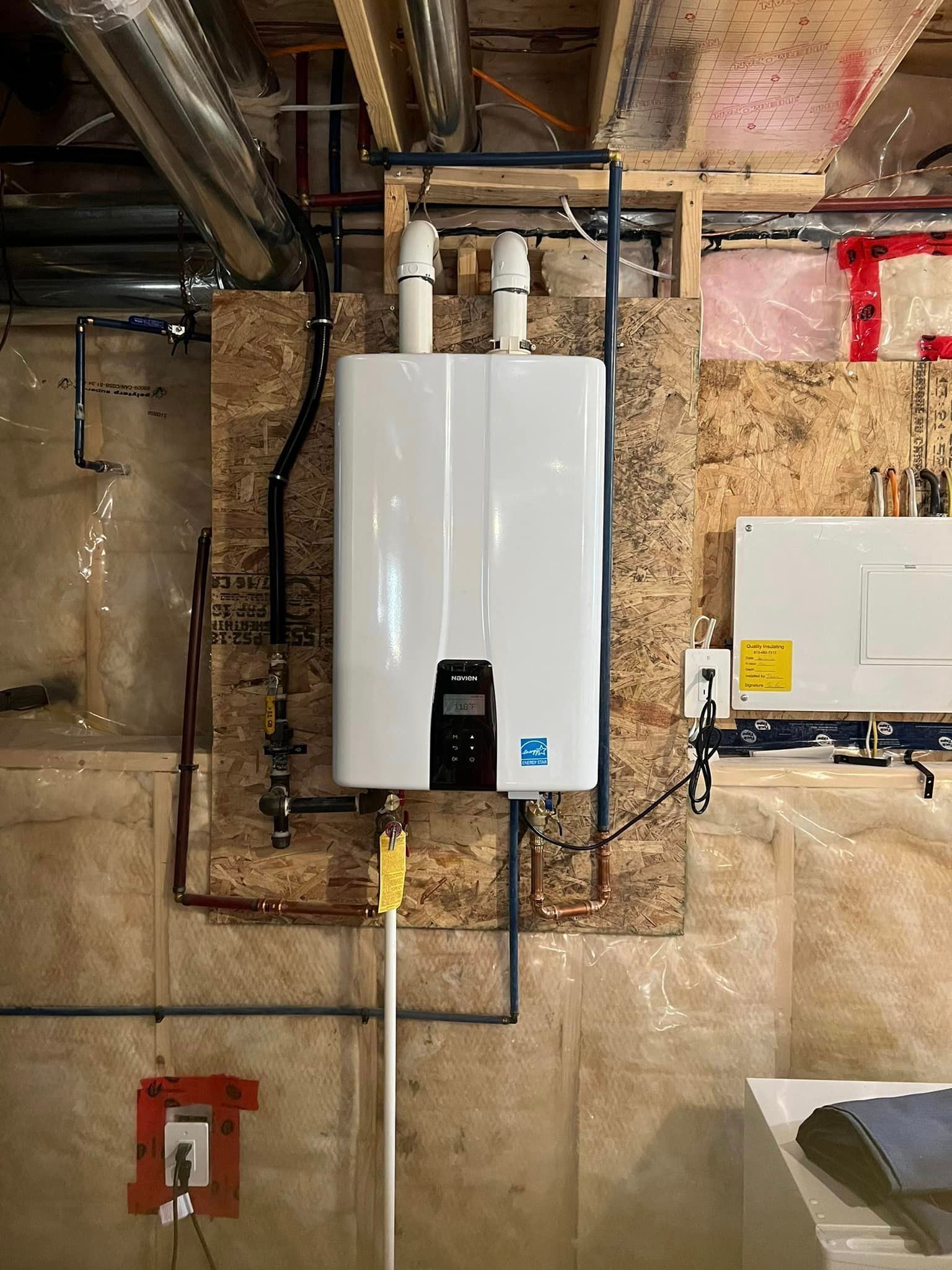 A white tankless water heater mounted on a wood panel in a basement, with black and copper pipes.