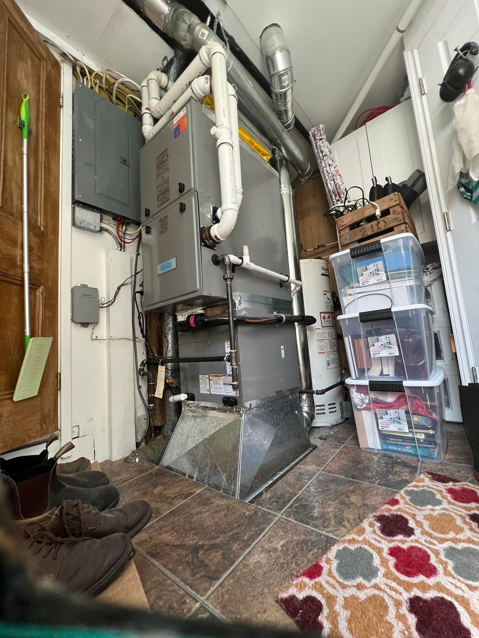 A furnace and storage containers in a cluttered room. The furnace is metallic gray. The floor has a rug.