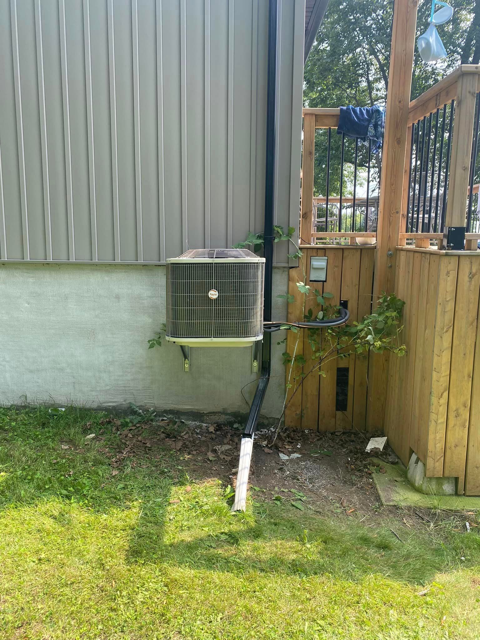 AC unit on a building exterior with a drain pipe, beside a wooden deck and grass.