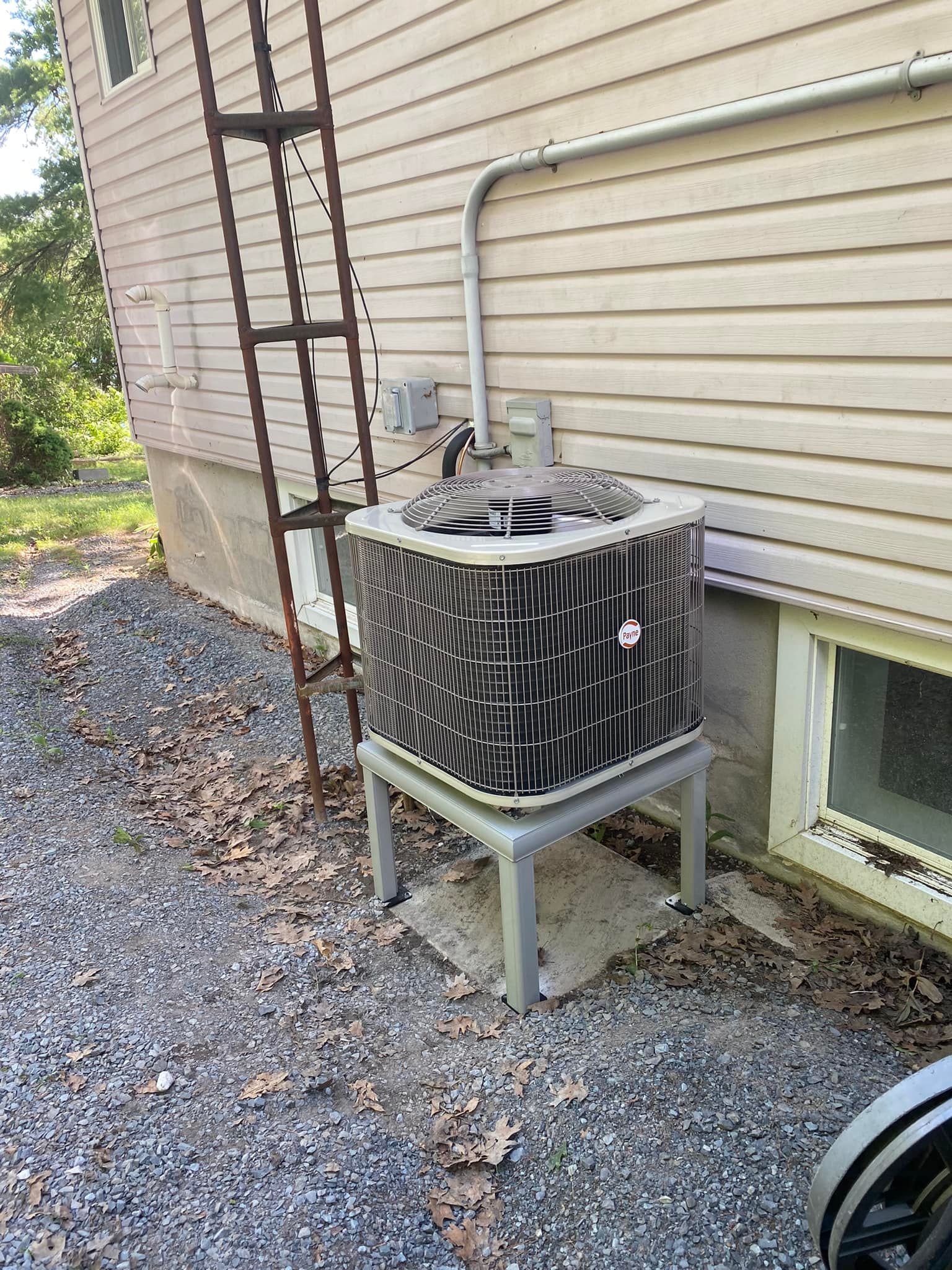 AC unit on a stand next to a house with a ladder. Gray gravel and fall leaves surround it.