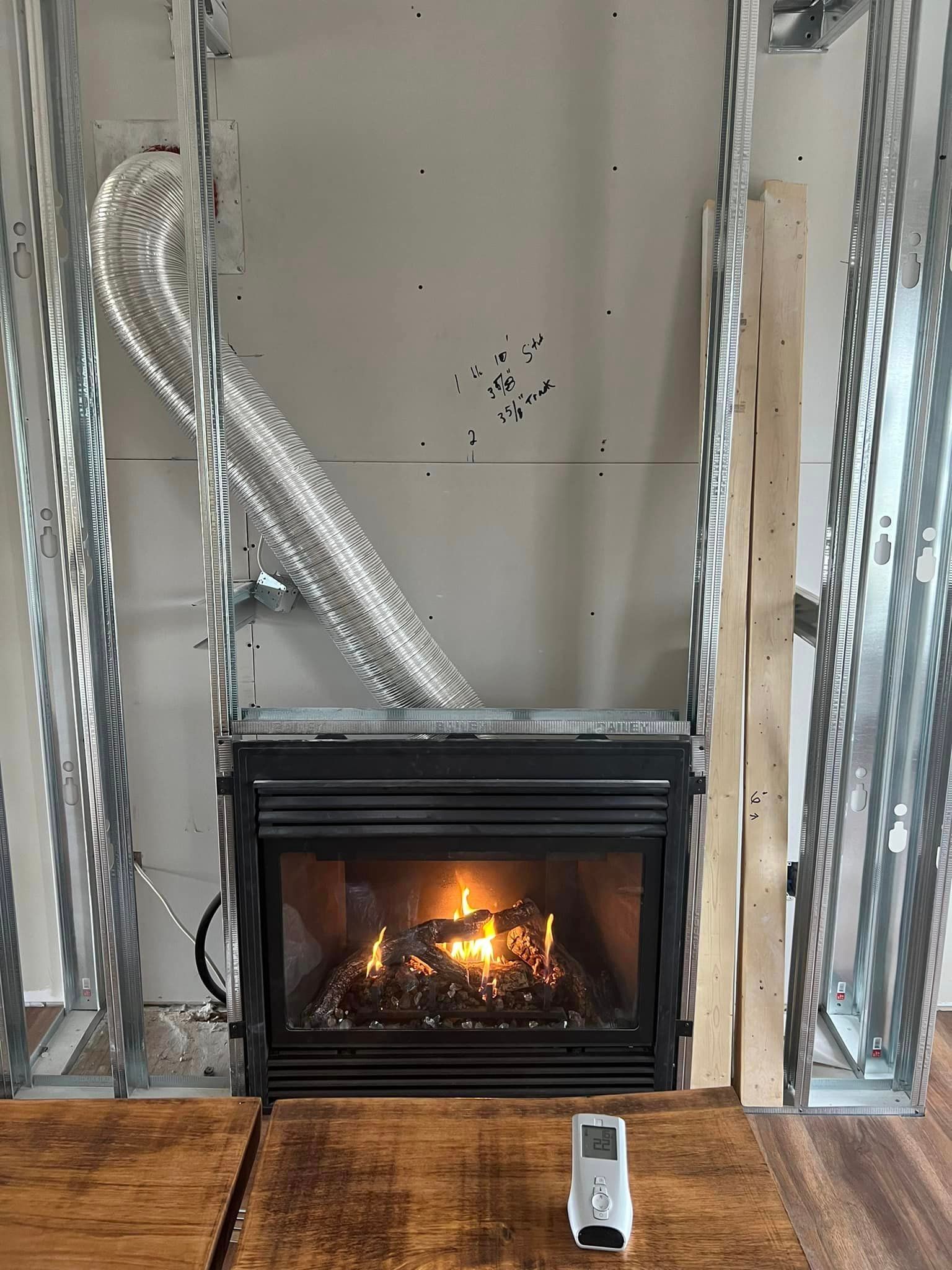 Fireplace under construction with burning flames, metal framing, and a remote on the wooden floor.