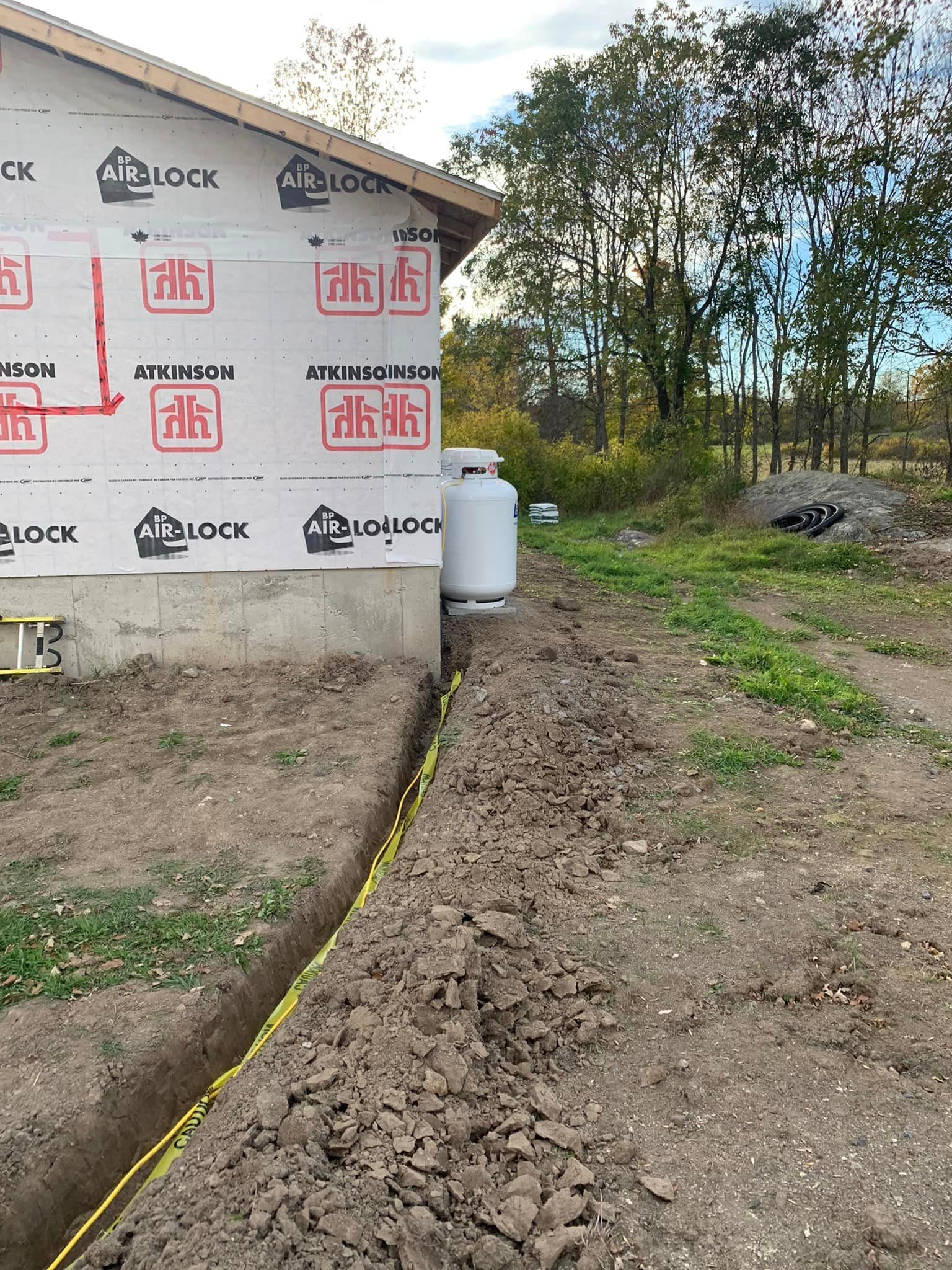 Trench with yellow caution tape running along the side of a house with a propane tank in the background.