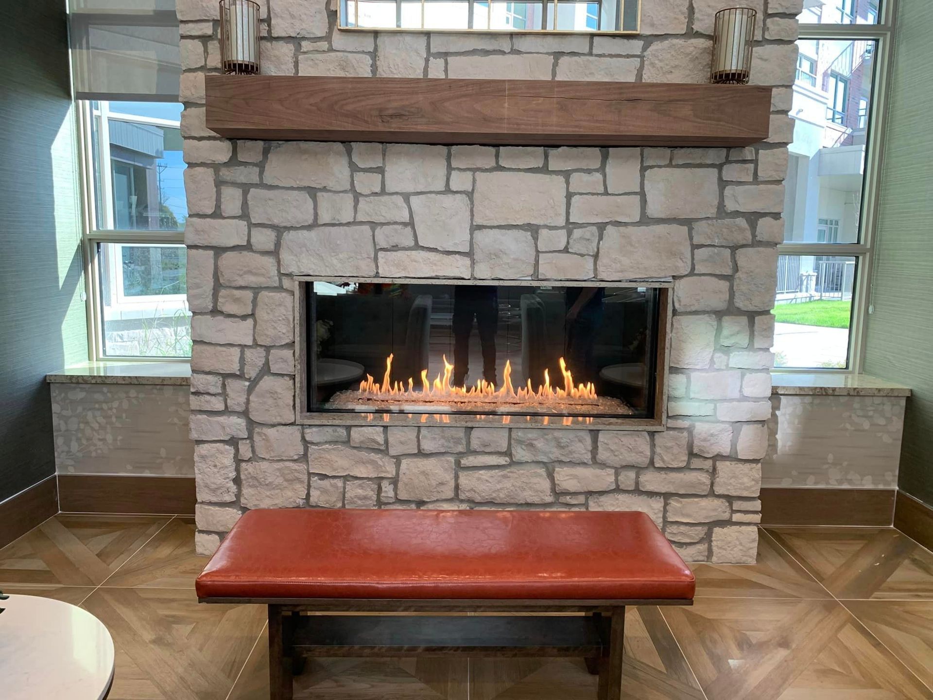 Stone fireplace with fire, wooden mantel, and leather bench in front. Windows on either side.