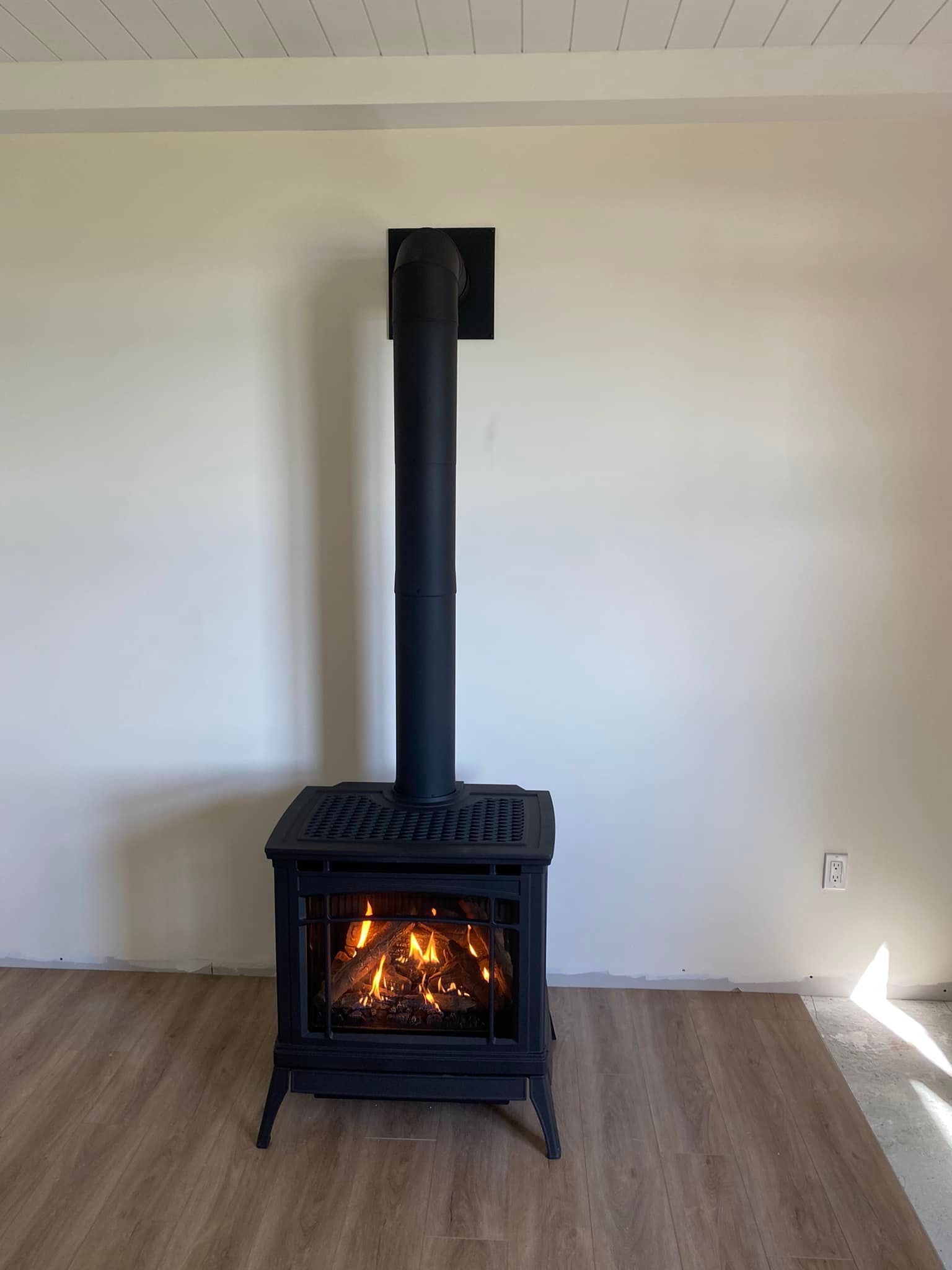 Black wood-burning stove with lit fire, black chimney, against white wall. Brown wood flooring.