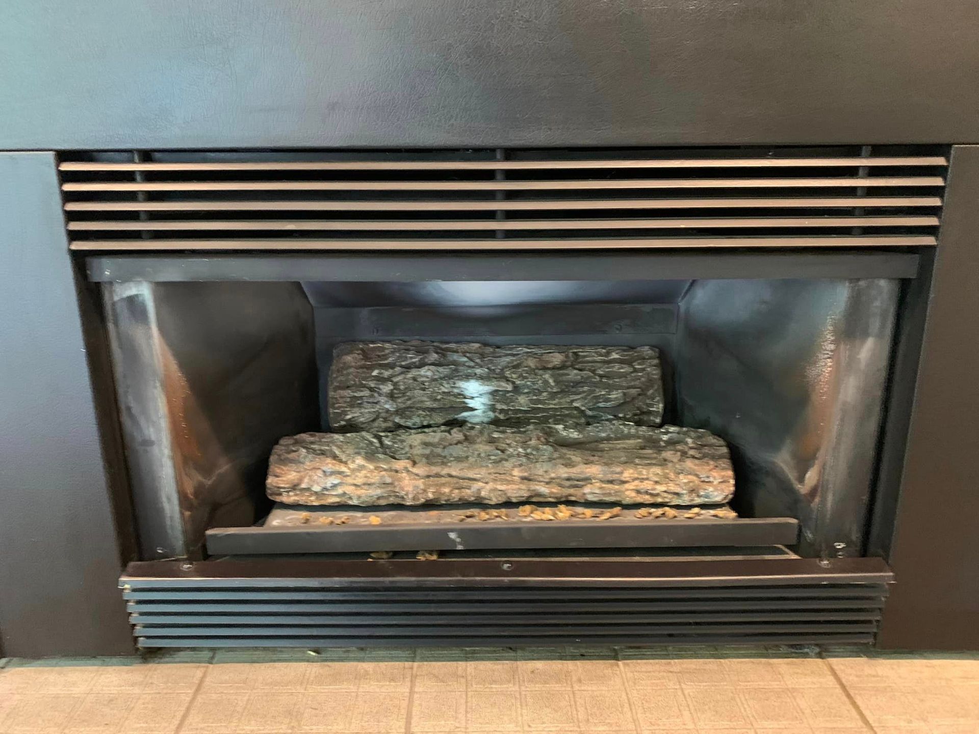 A gray fireplace with artificial logs inside. The fireplace has a black metal exterior and a vent at the top.