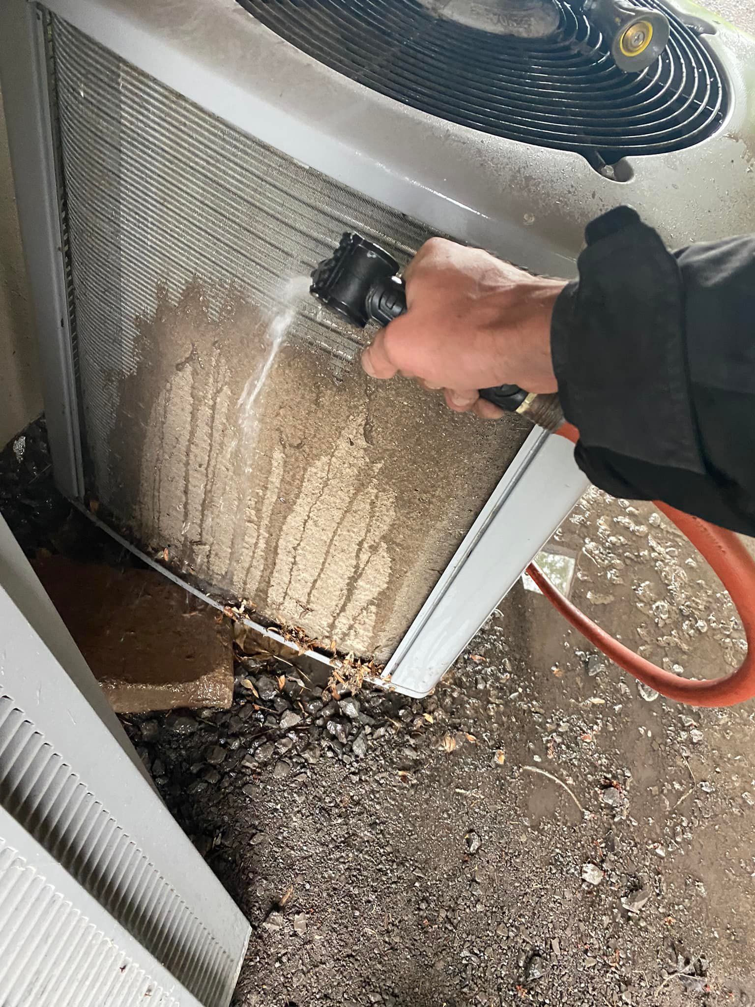 Person cleaning an air conditioner with a hose. The unit is covered in dirt, outside on gravel.