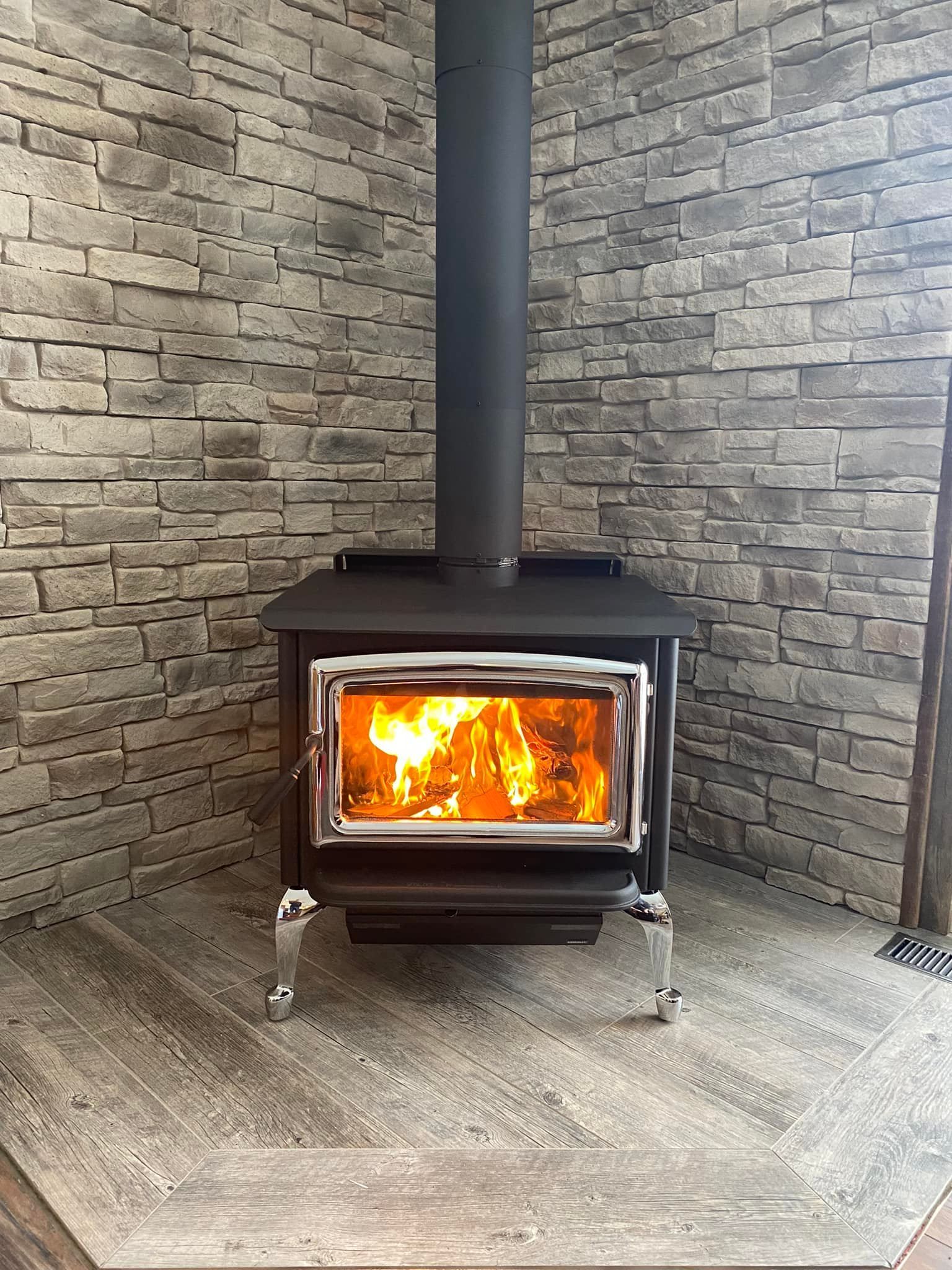 Wood-burning stove with flames, black metal, silver legs, in front of a stone wall.