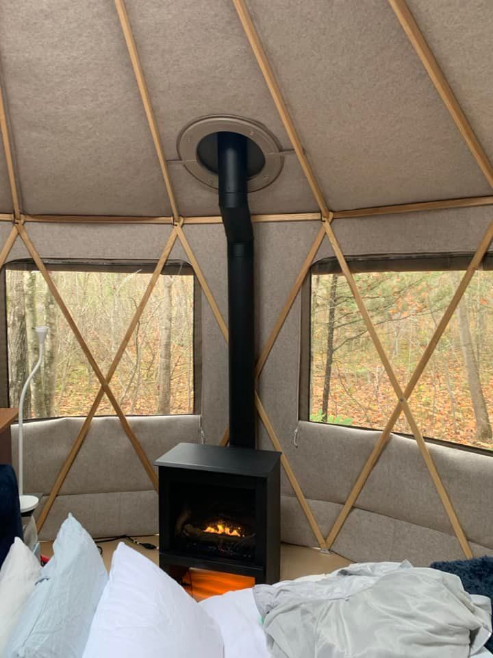 Cozy yurt interior with a black fireplace, windows, and wooden support beams.  Trees visible outside.