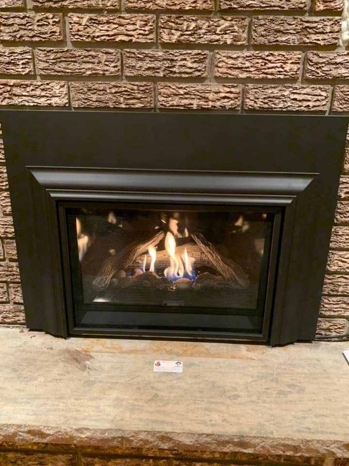 Black gas fireplace with lit flames, set against a brick wall and stone hearth.