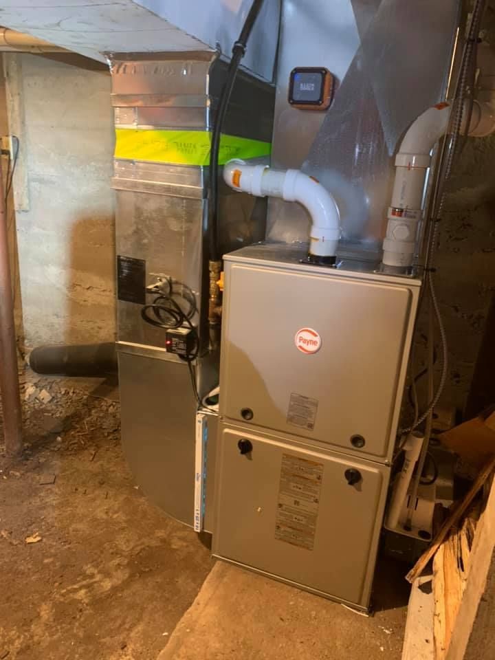 Furnace unit with ductwork in a basement. Gray metal components, white PVC pipes, and a concrete floor.