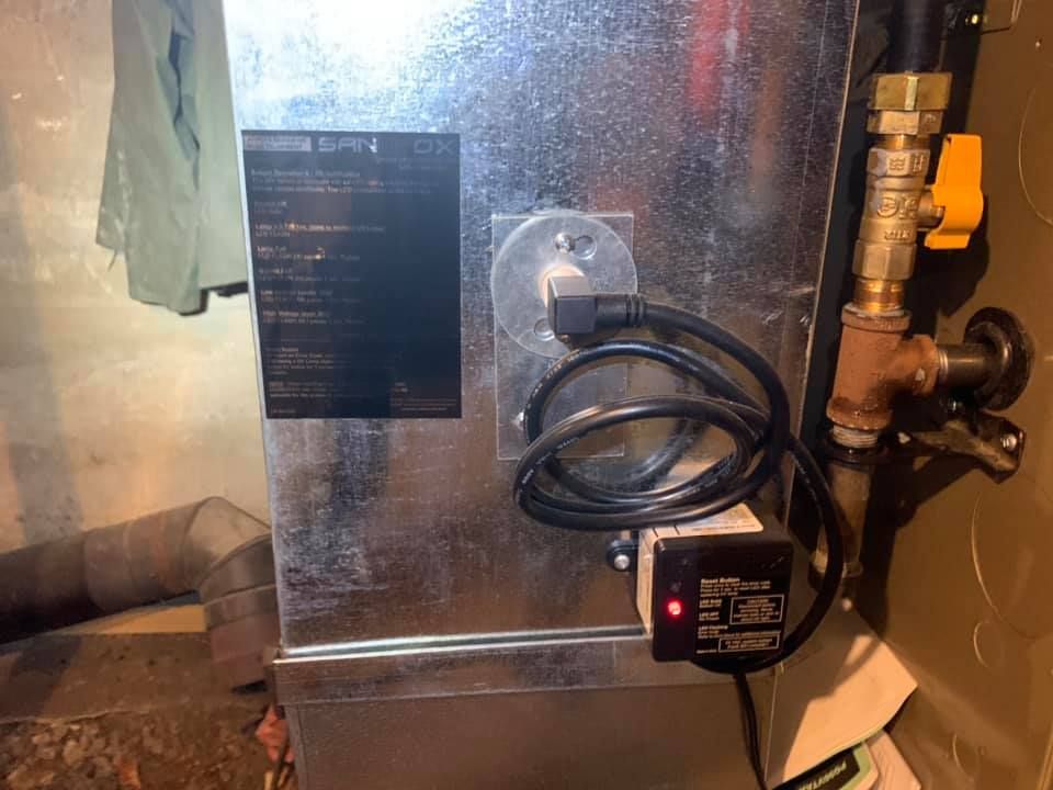 A furnace with a black power cord plugged in, a red light on a connected device, and gas pipes.