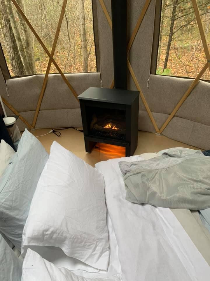Cozy interior of a yurt with a bed and electric fireplace; forested backdrop through windows.