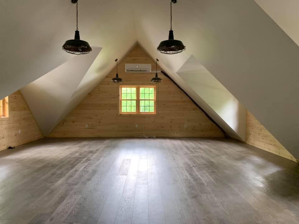 Spacious attic room with wood floors, sloped white ceilings, and exposed wooden wall with window and pendant lights.