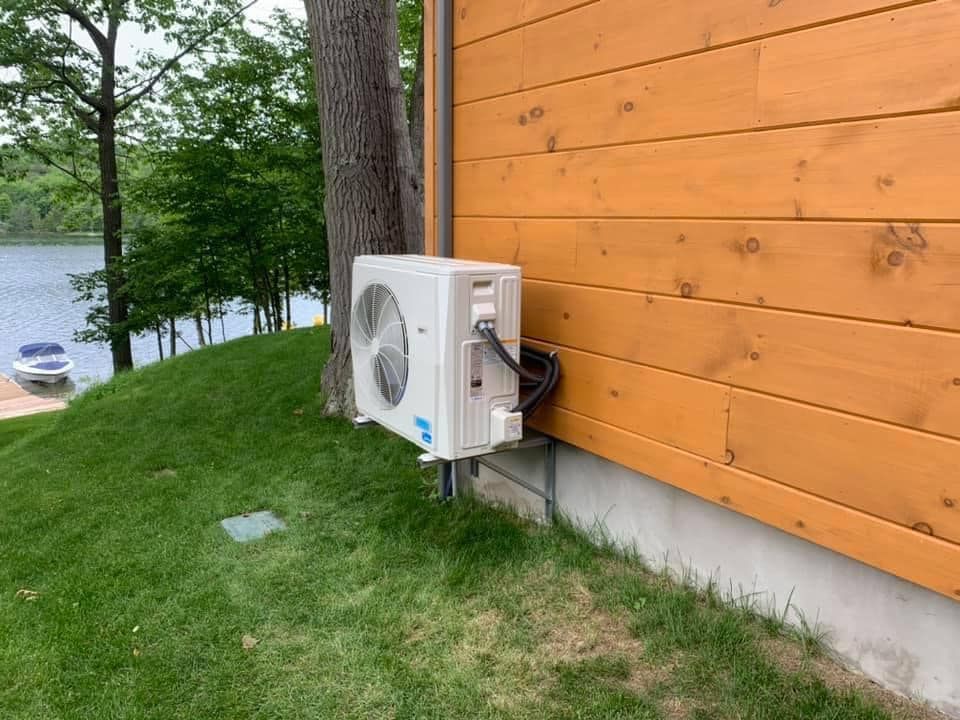 An outdoor air conditioning unit mounted on the wooden siding of a building next to a grassy hill and a lake.