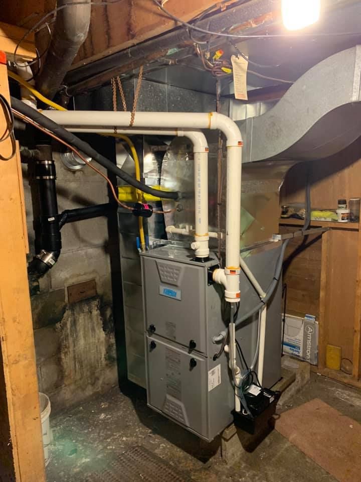Furnace and ventilation system in a basement, with pipes and ductwork. Grey and white, near a dark wall.