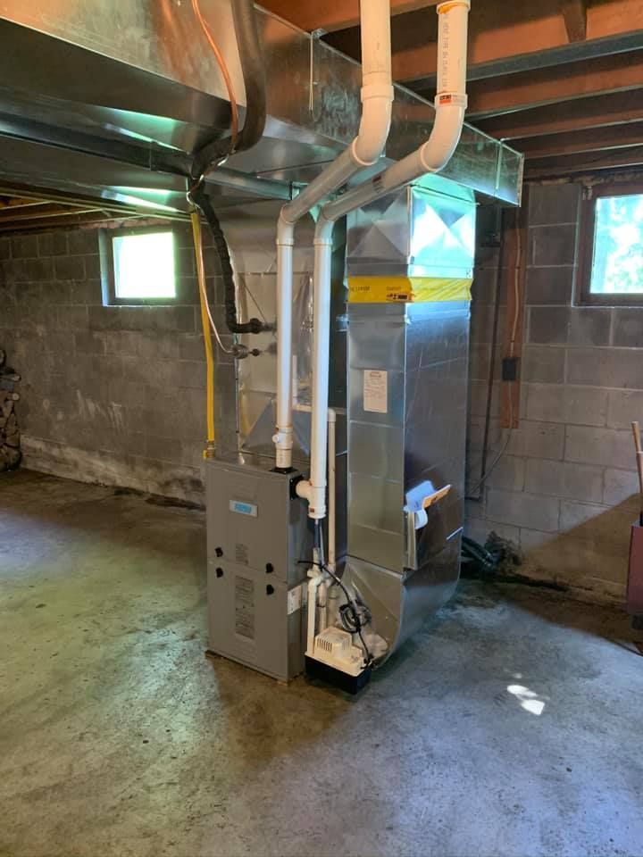 Furnace in a basement, with silver metal and white pipes. Cement block walls, gray floor.