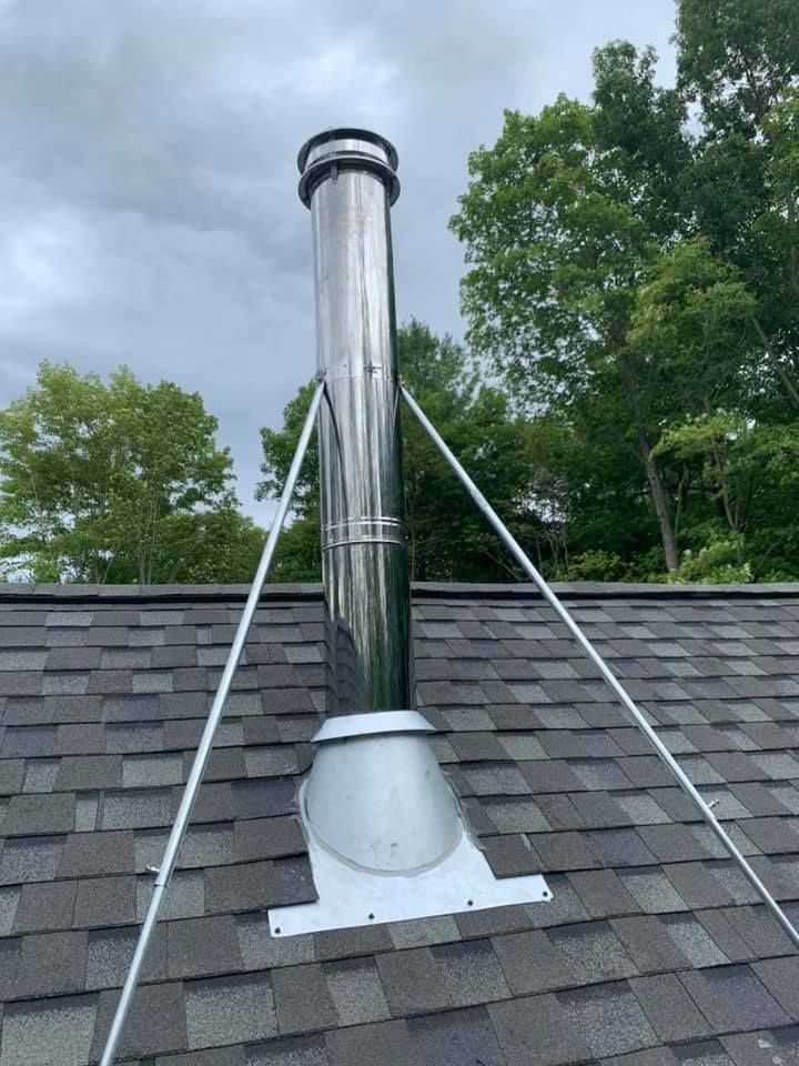 Shiny metal chimney pipe on a roof, supported by diagonal guy wires against a cloudy sky.