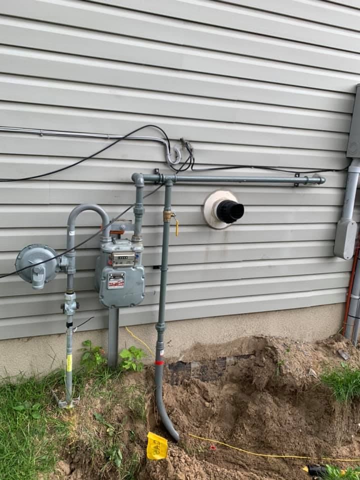 Gas meter and pipes attached to a light gray house. A hole is dug around the pipe near the ground.
