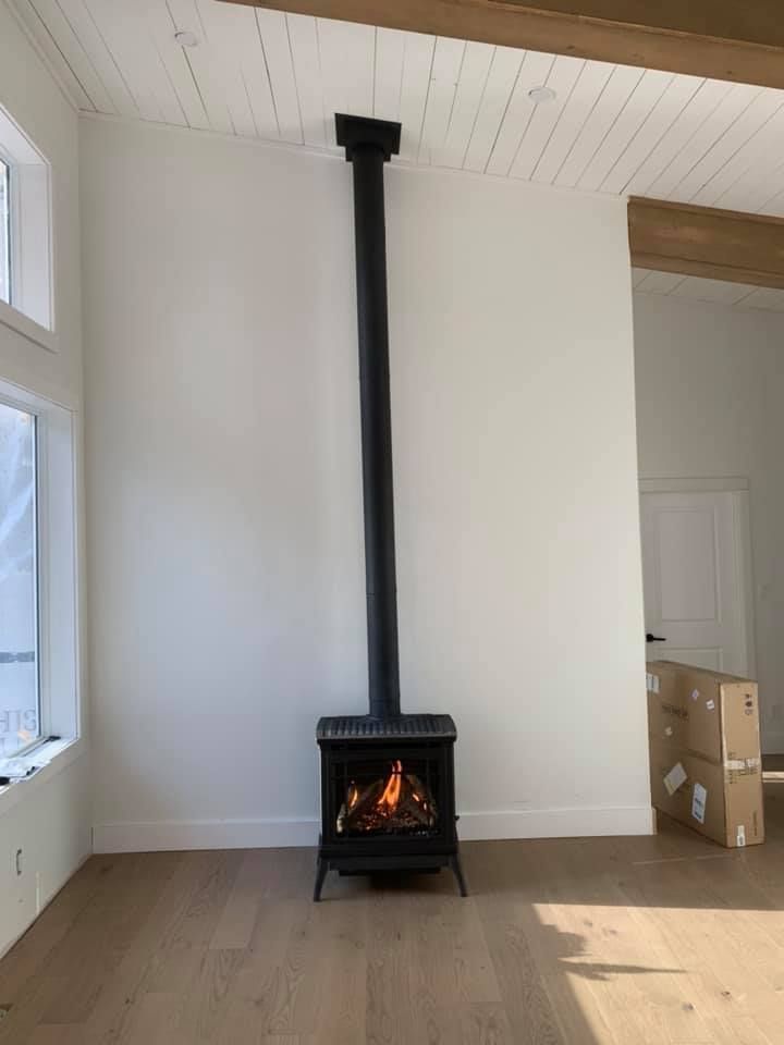 Black stove with lit fire and tall black chimney against a white wall, in a light-floored room.