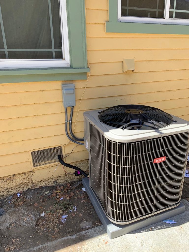Air conditioning unit outside a yellow house, near a window and electrical box.