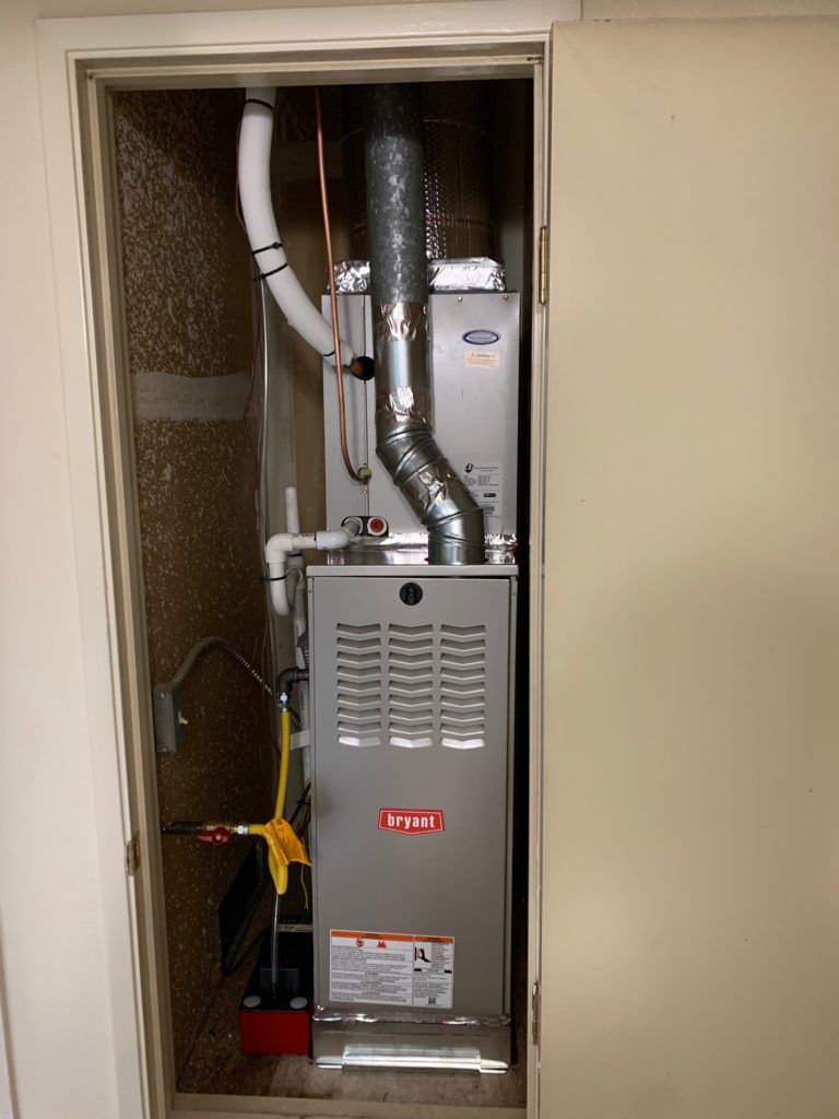 A silver Bryant furnace installed in a utility closet with a metal exhaust pipe and white piping.