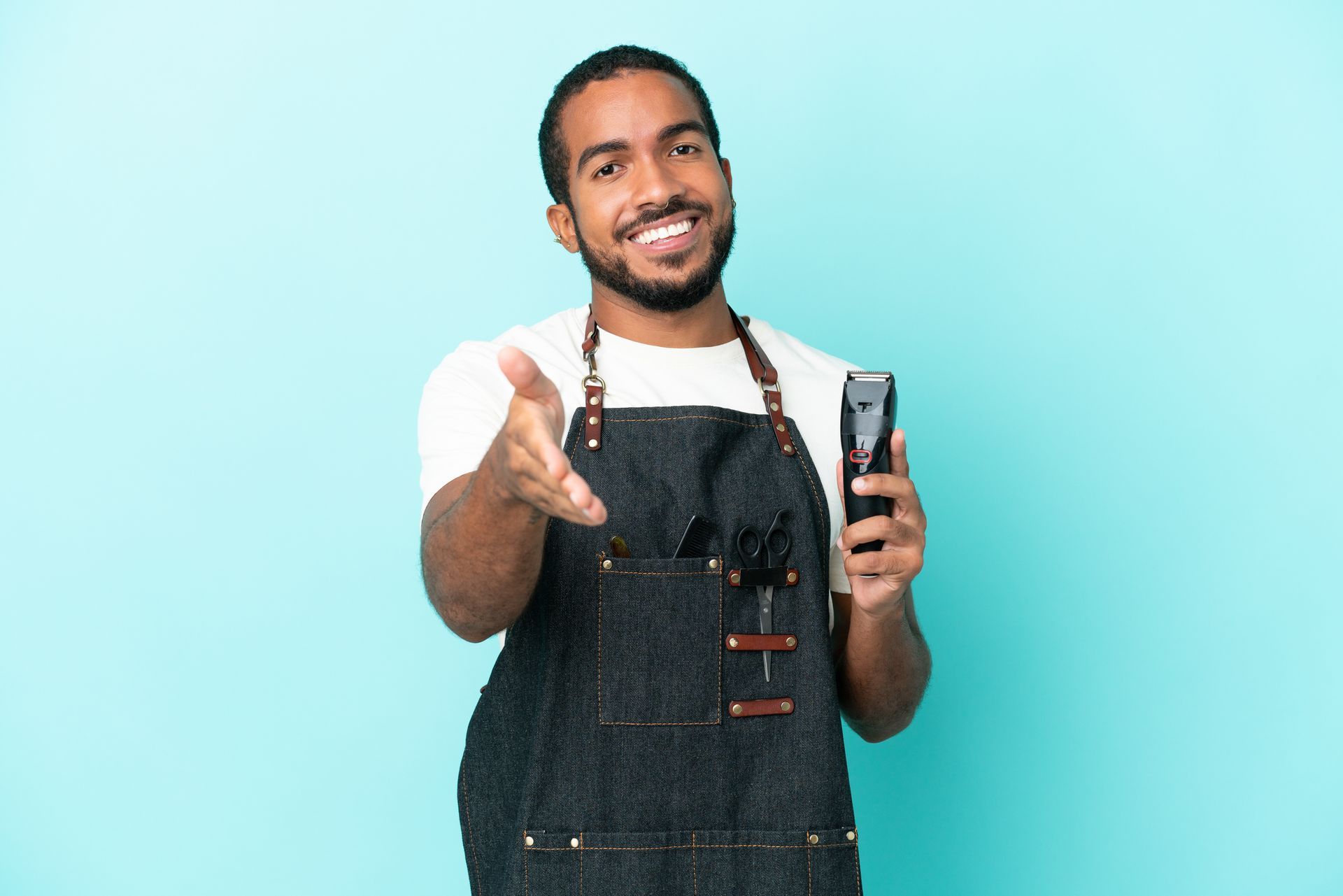 Barber with a welcoming hand, holding hair clippers, wearing apron, smiling.