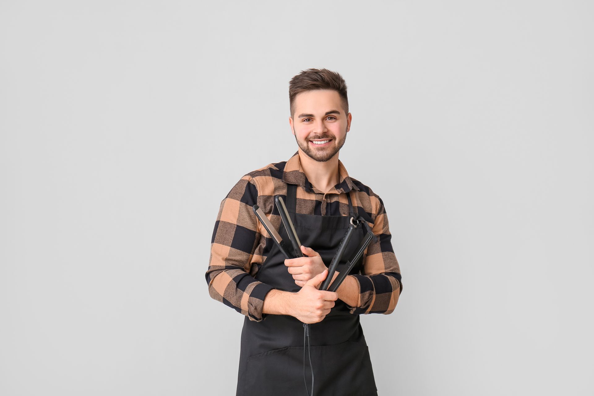 Man holding hair tools, wearing plaid shirt and black apron, smiling.