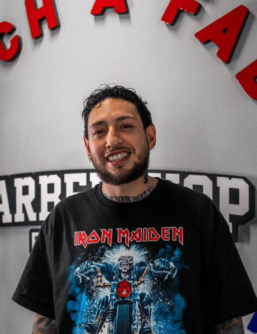 Hair stylist holding hairbrushes, smiling, wearing apron and plaid shirt.