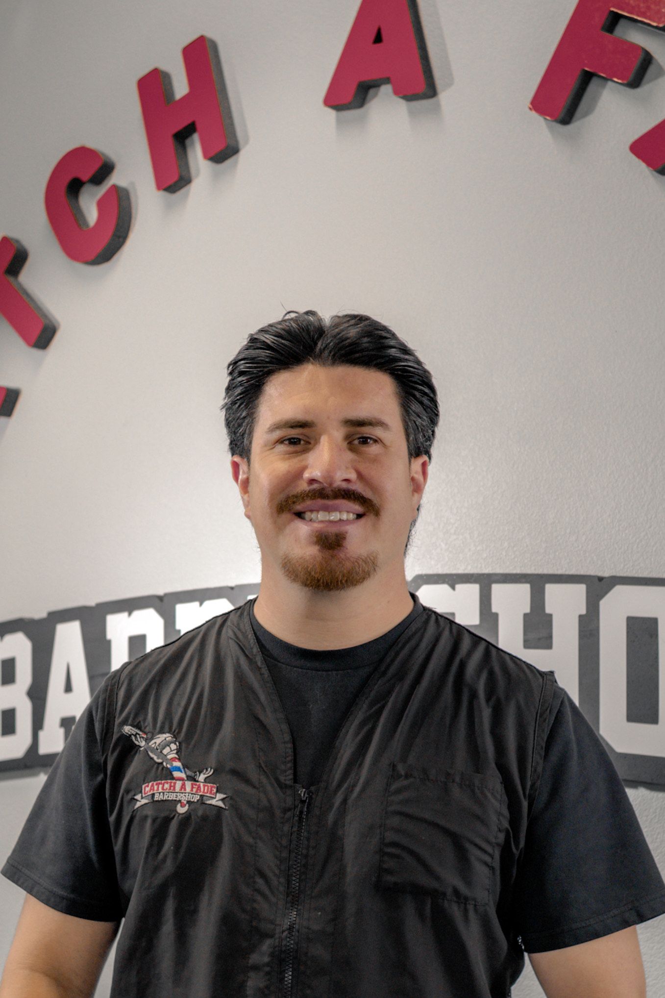 Man with red beard and tattoos smiles, points forward, wearing an apron over a gray shirt.