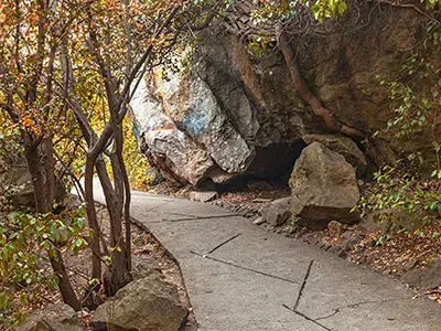 A paved path curves past large boulders and a rock overhang. Trees and foliage frame the scene.
