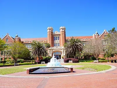 Building with brick facade, two towers, fountain in foreground, palm trees, sunny day.