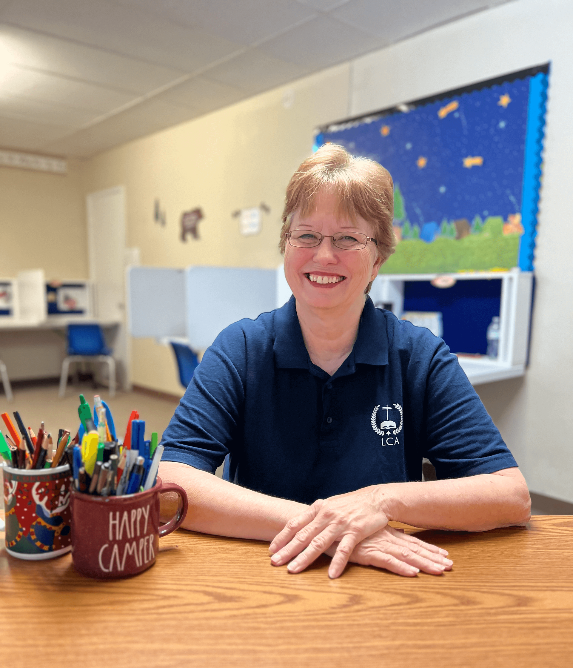 A woman is sitting at a table with a cup of pencils on it.