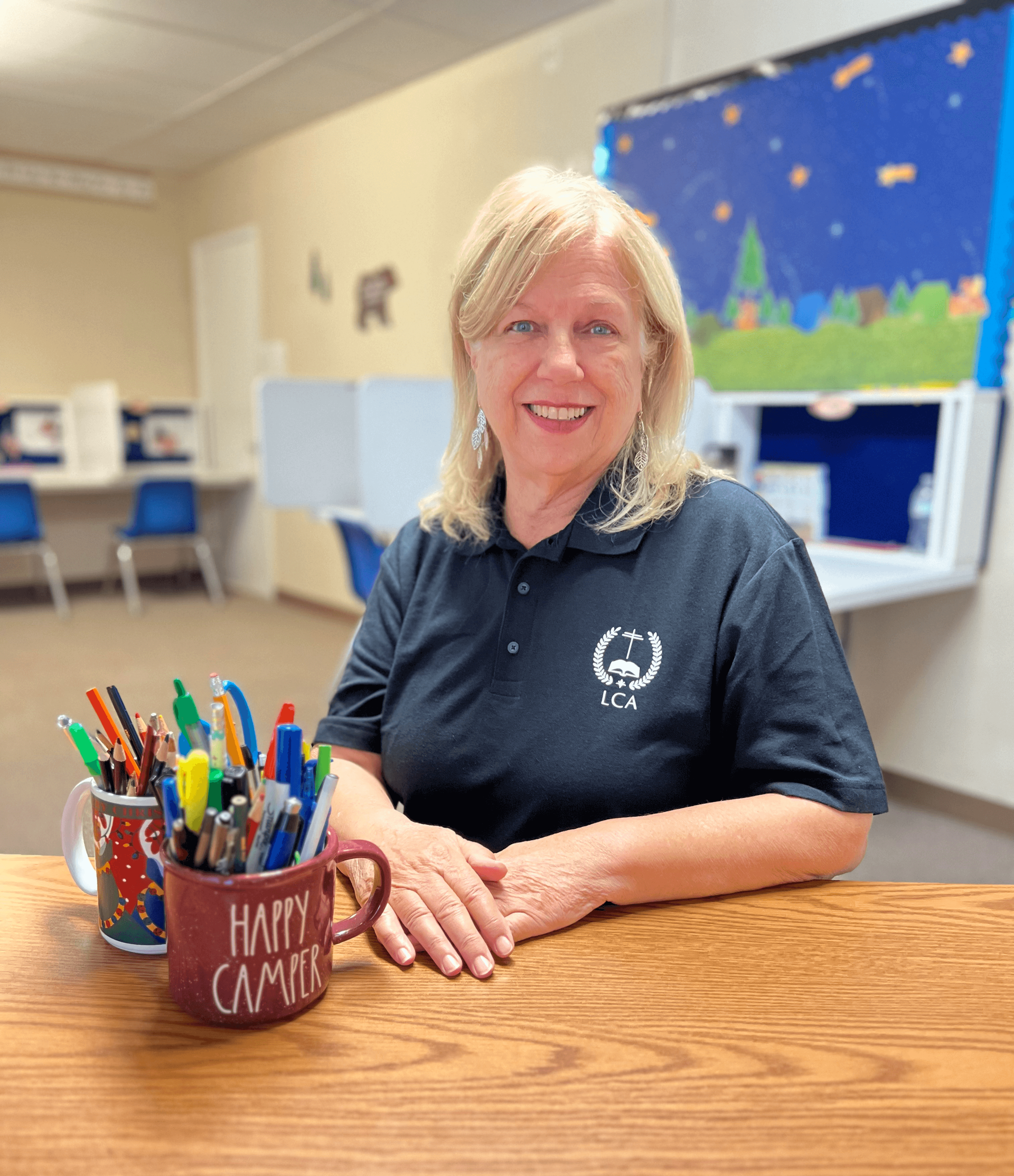 A woman is sitting at a table with a cup of pencils in front of her.