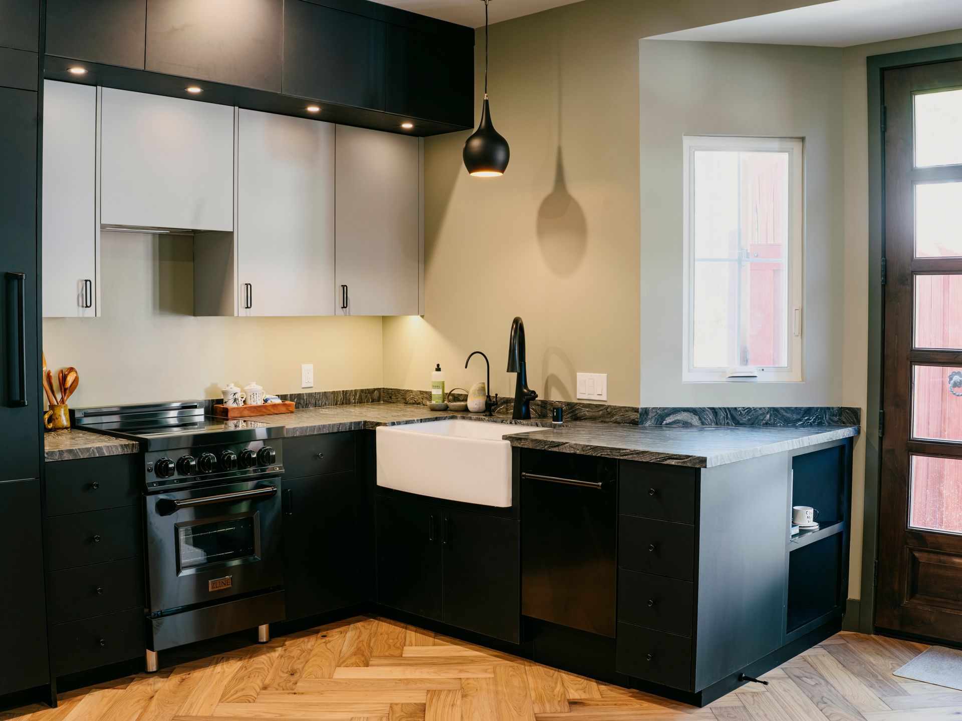 Modern kitchen with black and gray cabinets, stainless steel appliances, and herringbone wood floors.