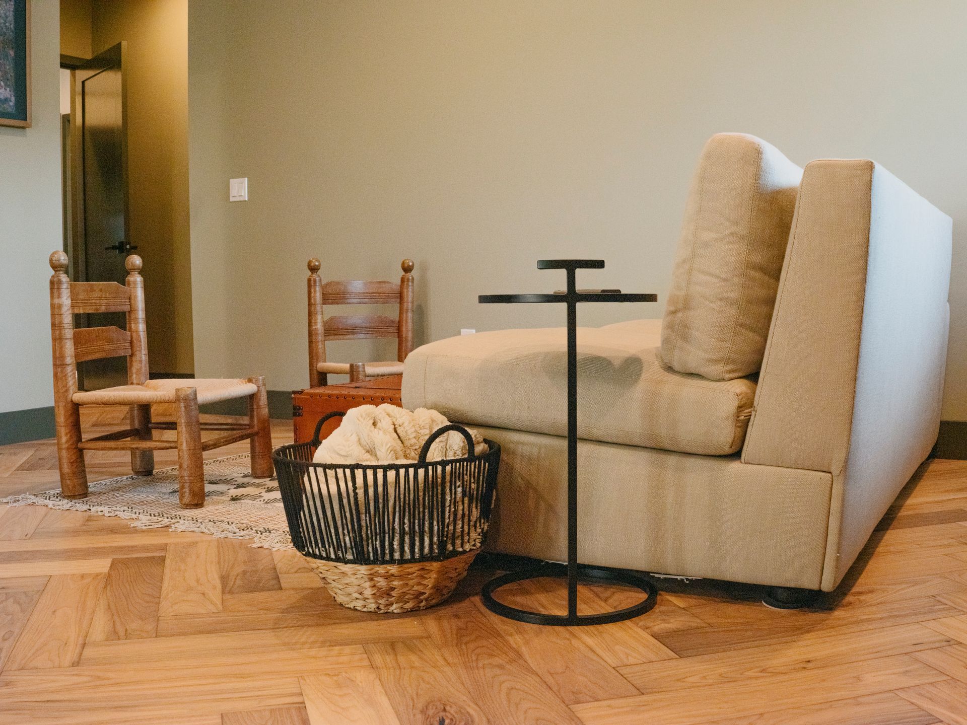 A tan couch, basket, and side table in a room with wooden floors. Two wooden chairs are in the background.