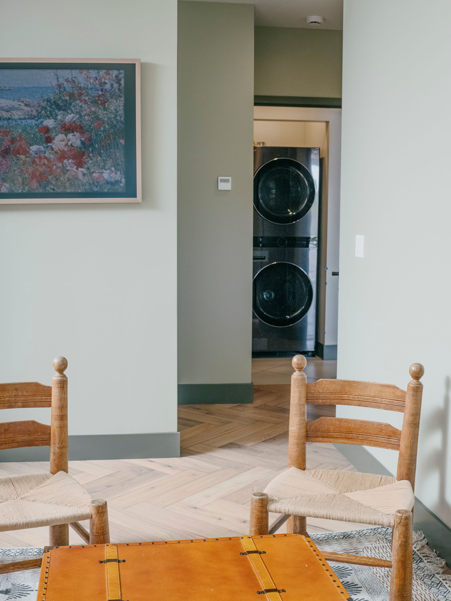 Two chairs, a trunk, and a painting frame a hallway leading to a stacked washer and dryer.