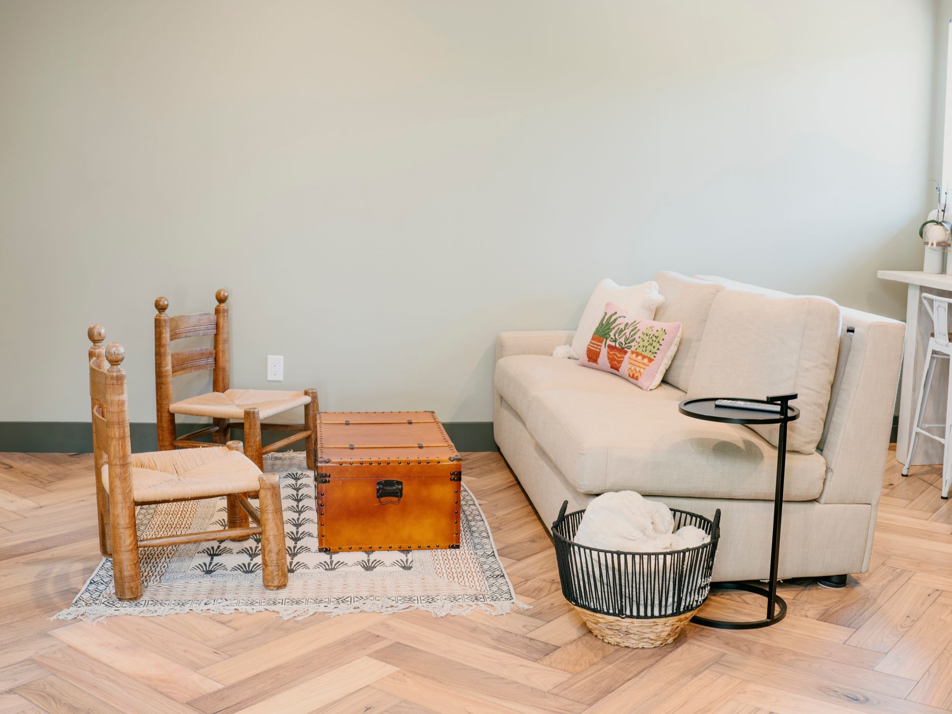 Living room: couch, chairs, trunk, rug, side table, basket, light green wall, wood floor.