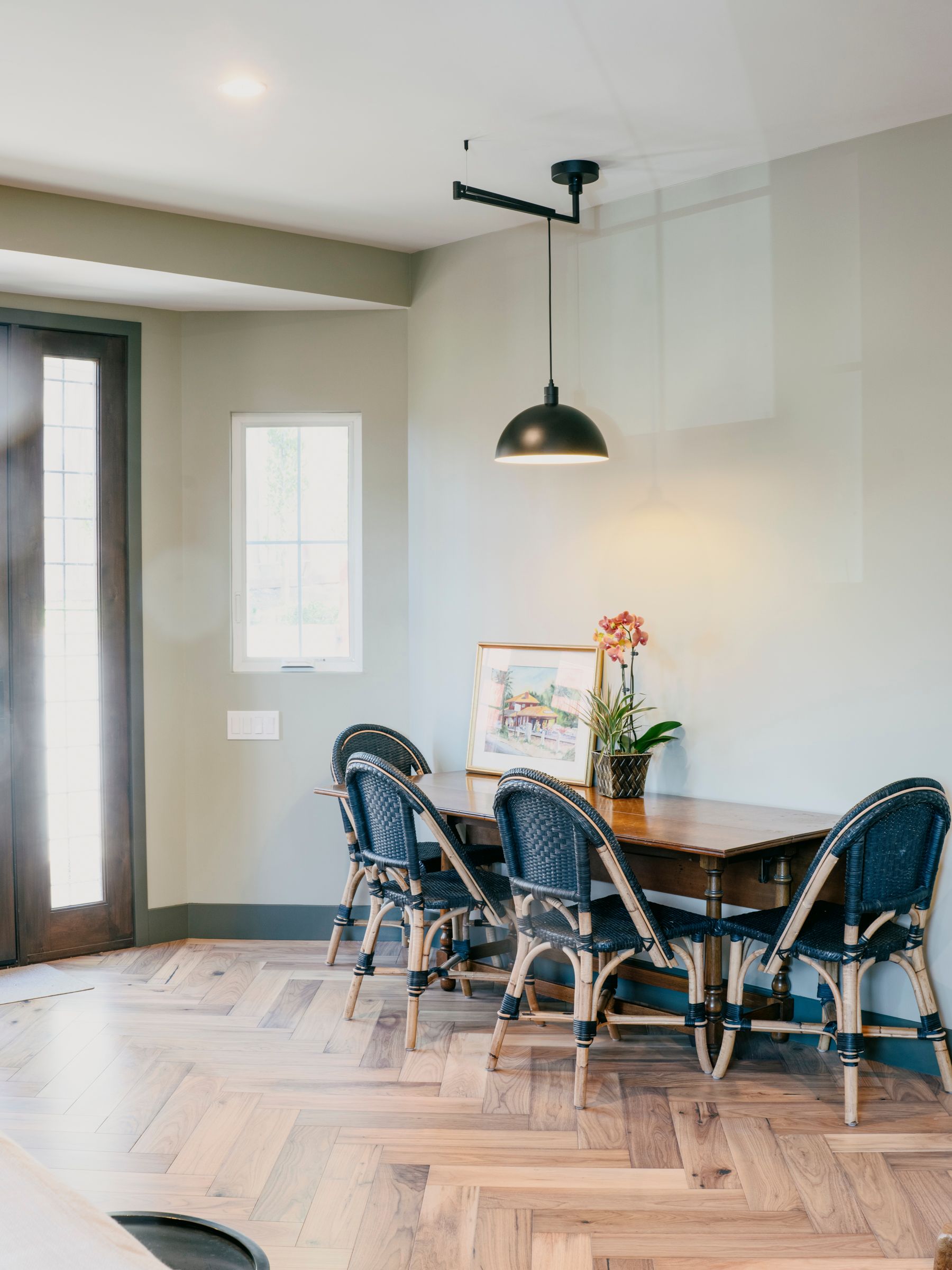 Dining room with wooden table, rattan chairs, and herringbone floors. Black pendant light hangs above.