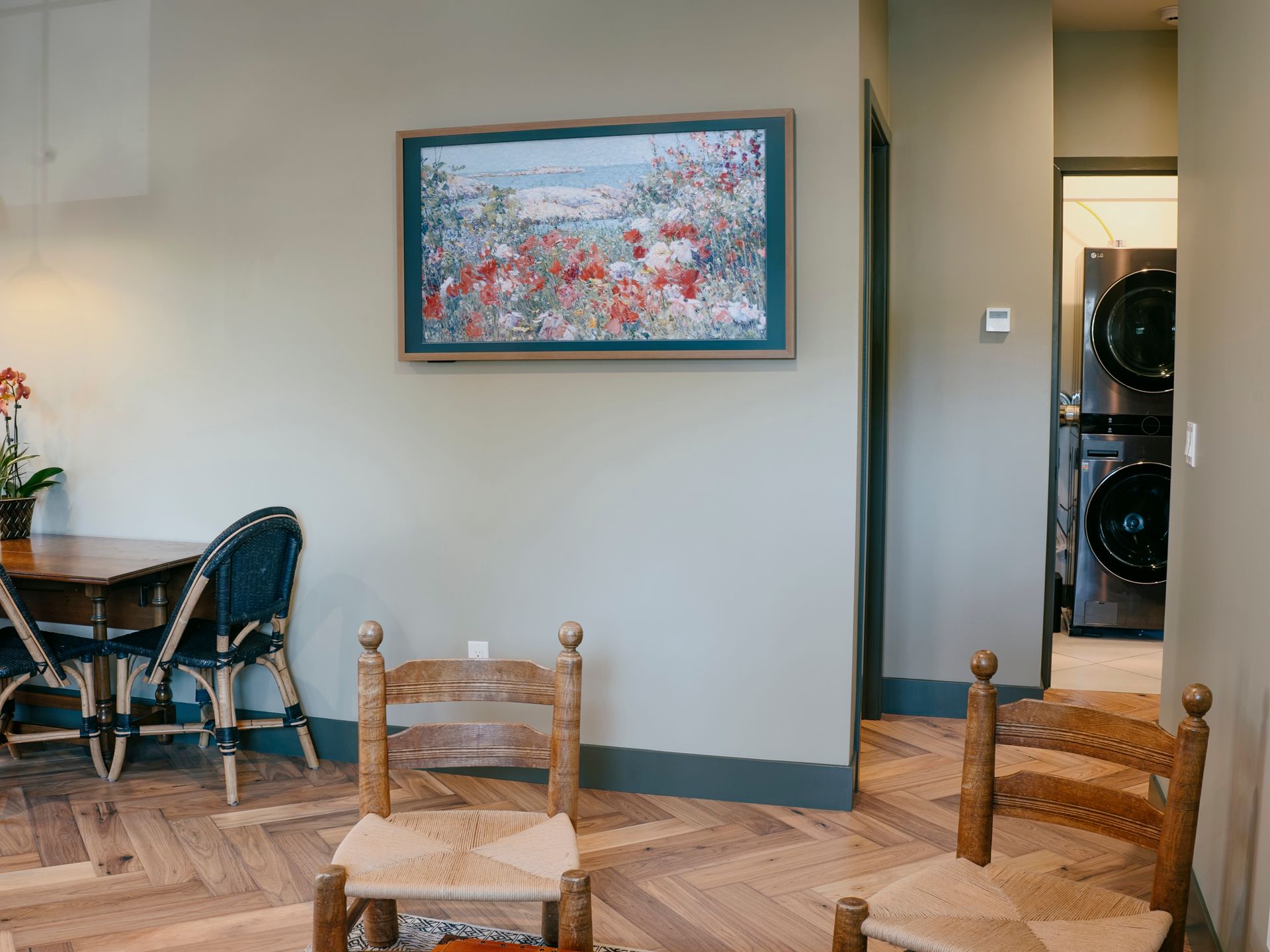 A living room with wooden chairs, a table, and a painting. Laundry machines are visible in the hallway.