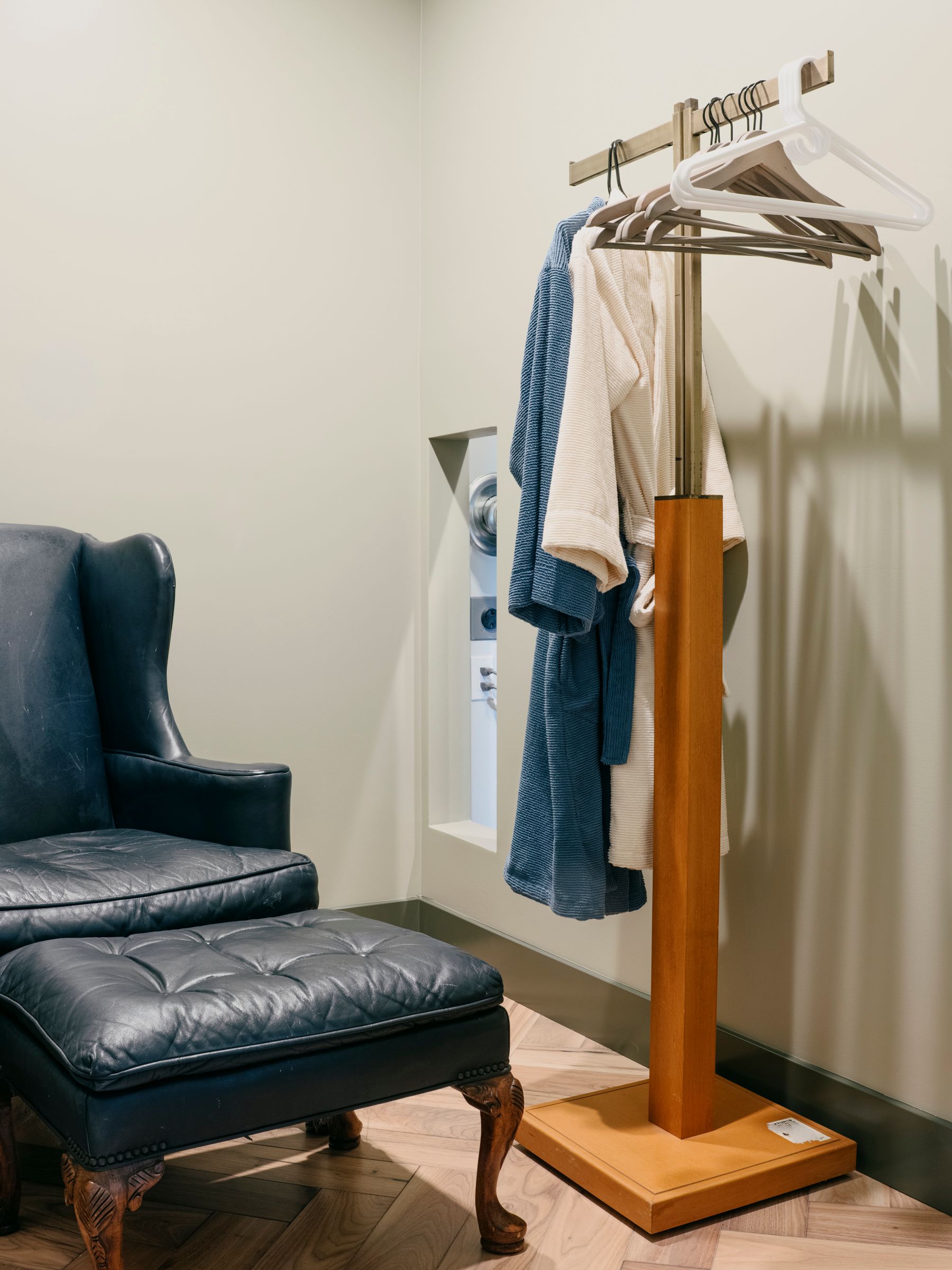 Blue armchair and ottoman next to a robe stand with robes hanging in a neutral-toned room.