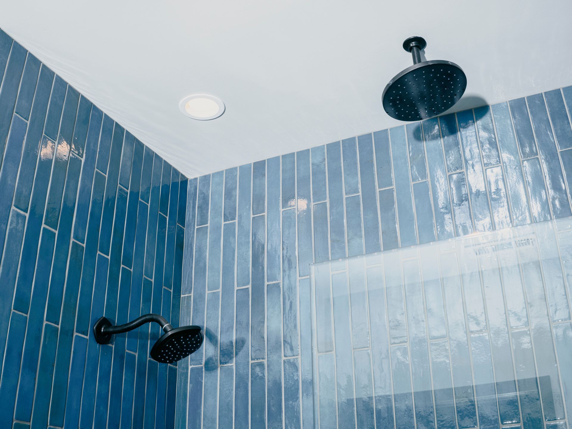 Blue tiled shower with two black showerheads and a recessed ceiling light.