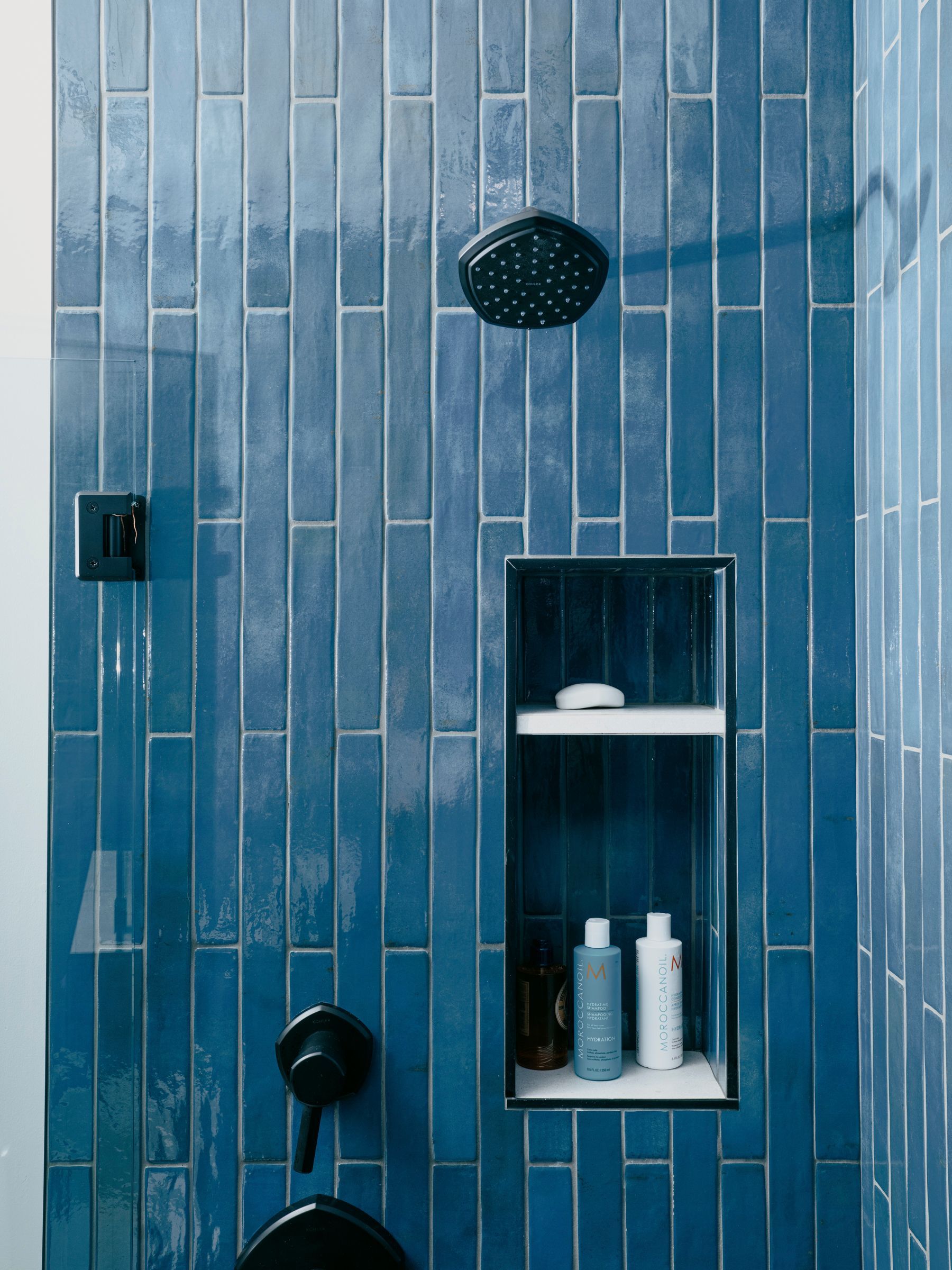 Blue tiled shower with black fixtures, a shower niche with toiletries, and a black shower head.