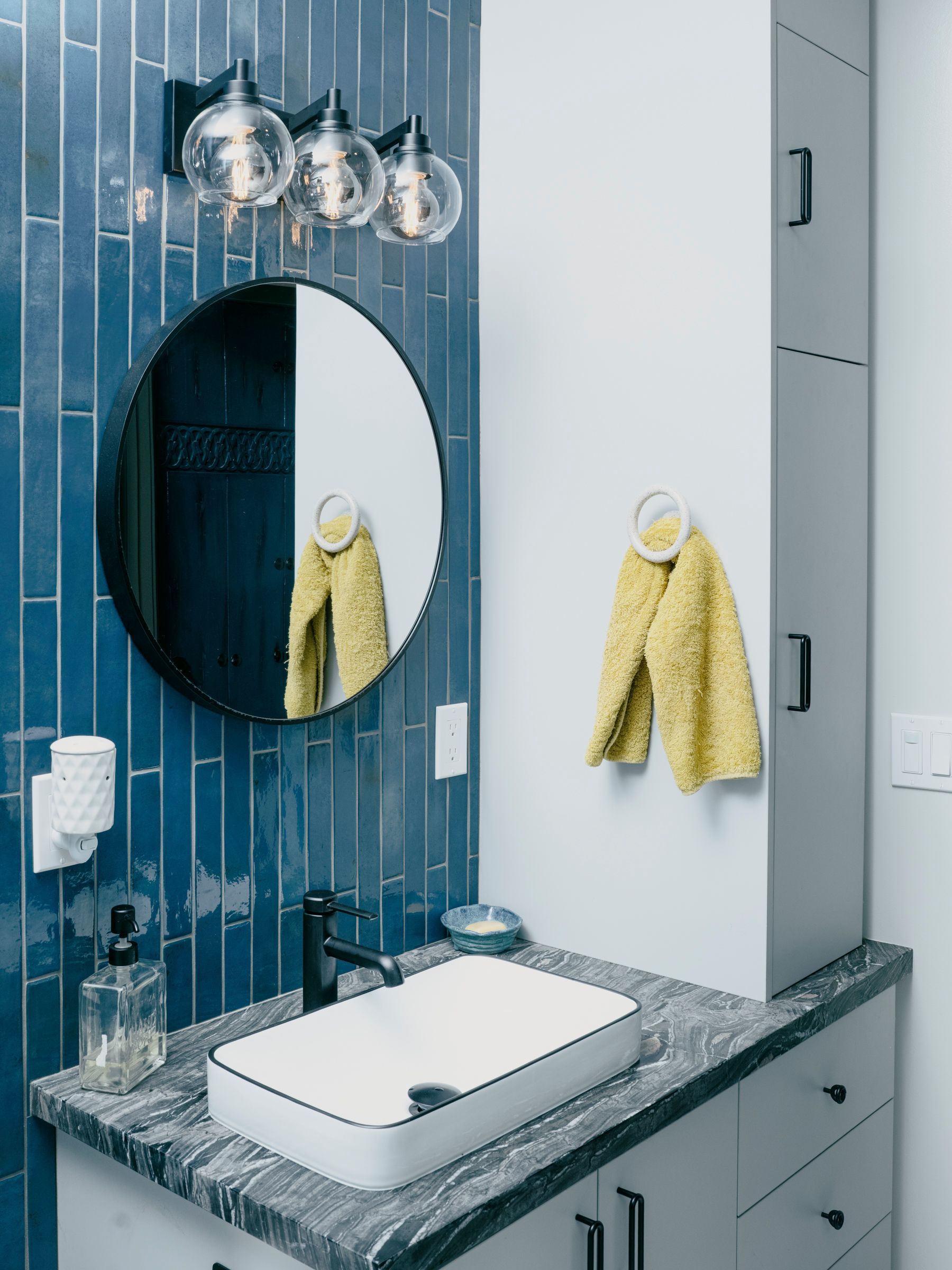 Bathroom with blue tiled accent wall, round mirror, and floating sink. White countertop, and gray storage cabinet.